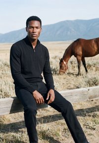Homme en tenue noire assis sur une clôture en bois dans un champ sec avec un cheval brun qui broute à proximité et des montagnes en arrière-plan.