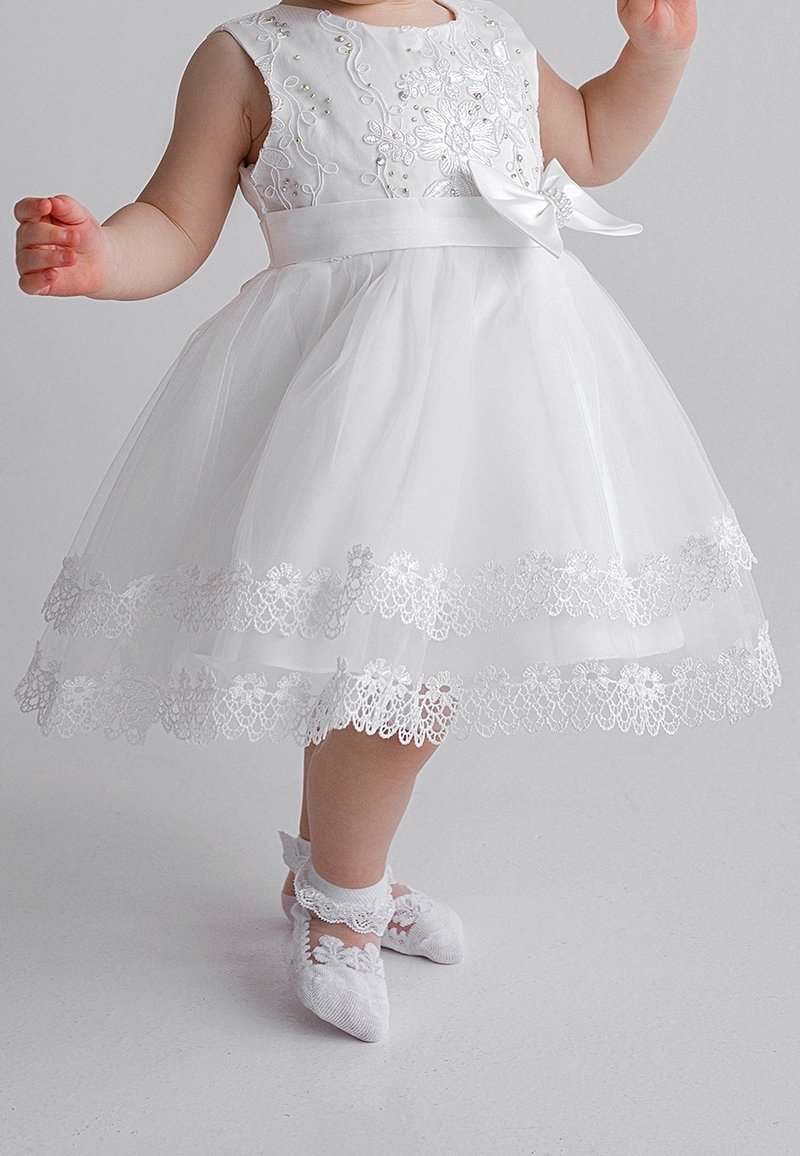 White dress with floral embroidery, a satin bow at the waist, and a layered tulle skirt with lace trim. White decorative socks with lace accents.