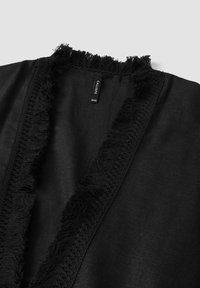 Czarny materiał odzieżowy z frędzlami na dekolcie i ozdobnym szydełkowym wykończeniem na brzegach. Metka pokazuje nazwę marki "CALLIOPE" oraz rozmiar "JEDNY ROZMIAR."