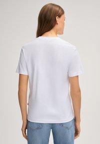 Weißes T-Shirt aus Baumwolle mit kurzen Ärmeln, Rundhalsausschnitt und geradem Schnitt. Hat ein kleines rotes Etikett auf der linken Seite nahe dem Saum.