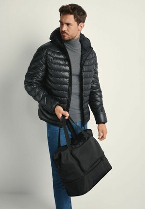 Czarna puchowa kurtka z kapturem, posiadająca pikowaną fakturę i zamek z przodu. Ciemnoszary golf pod spodem. Czarna torba sportowa trzymana w ręku.