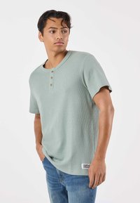 Ljusgröna kortärmade henley-tröja med tre bruna knappar, strukturerat tyg och en etikett vid fållen, kombinerad med blå jeans.