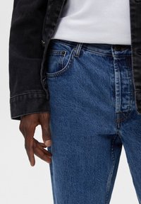 Vaqueros azules de mezclilla con un corte clásico, con cierre de botones, presillas para cinturón y detalles sutiles de costura, combinados con una chaqueta negra.