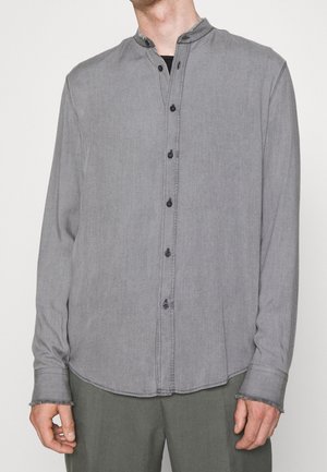 Man wearing a long-sleeve gray button-up shirt with a mandarin collar and dark pants, standing against a plain background.