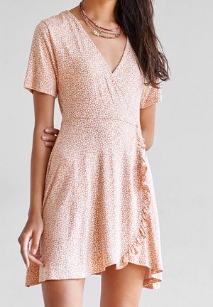 Femme portant une robe portefeuille à manches courtes rose pâle à motifs floraux avec bordure volantée et colliers perlés superposés, debout sur un fond uni.