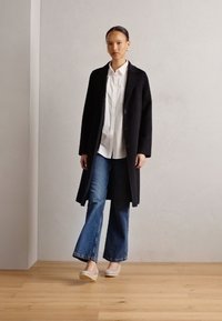 Long black coat over a white button-up shirt, paired with flared blue jeans and light-colored shoes; features clean lines and a minimal design.