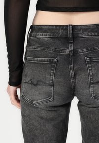 Grijze denim jeans met een vervaagde afwerking, voorzien van twee achterzakken en een hoge tailleband, gefotografeerd van achteren met een doorschijnend zwart topje zichtbaar.