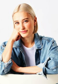 Modella che indossa una giacca di jeans azzurro chiaro sopra un top bianco a coste, con capelli lisci, corti e biondo platino e piccoli orecchini a cerchio in oro.