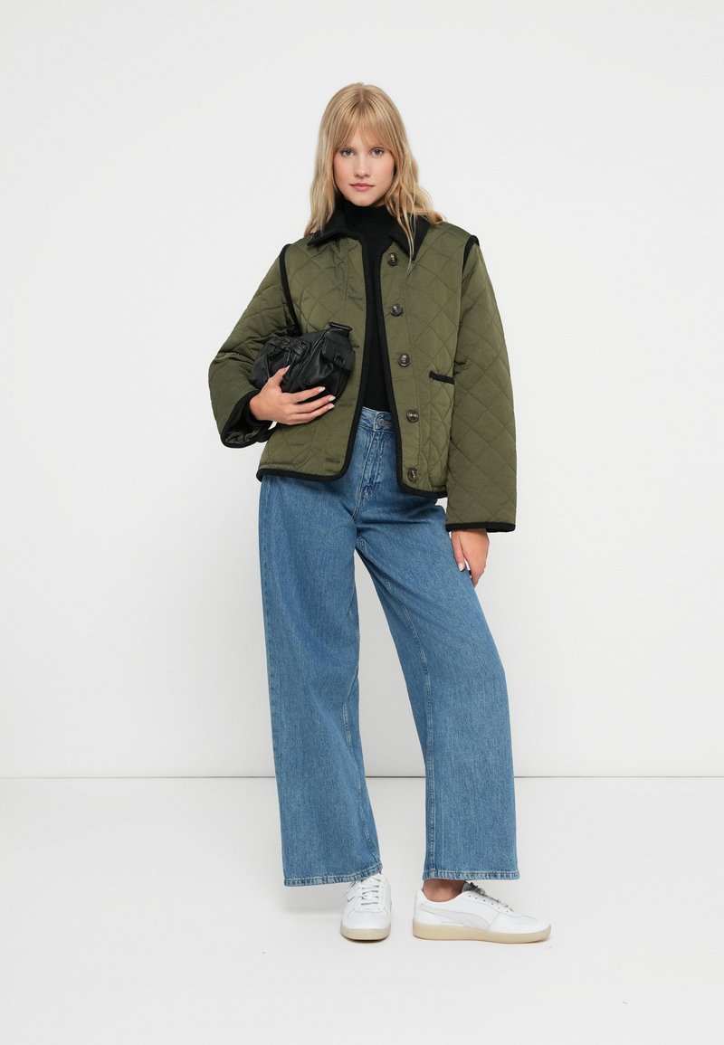 Olivgrüne gesteppte Jacke mit schwarzem Besatz und Knopfverschluss, getragen über einem schwarzen Rollkragenpullover, kombiniert mit hellblauen weit geschnittenen Jeans und weißen Sneakers.