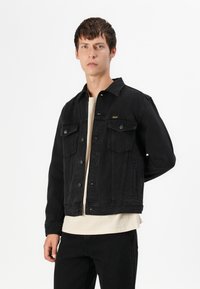 Black denim jacket with two chest pockets and snap buttons. Worn over a light beige shirt, featuring a classic collar and cropped design.