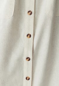 Beežist linast särk, millel on nööpidega esitasku ja kuus pruuni nööpi, samuti kaks rinnataskut. Kangas on tekstureeritud pinnaga.