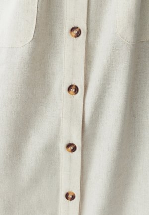 Světle béžová lněná košile s zapínáním na knoflíky, která má šest hnědých knoflíků a dvě náprsní kapsy. Tkanina má texturovaný povrch.