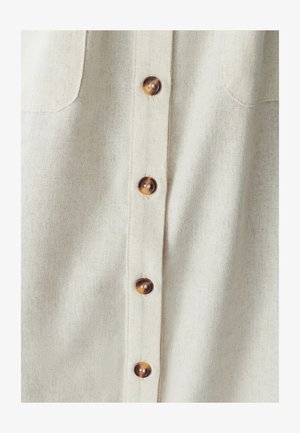 Světle béžová lněná košile s zapínáním na knoflíky, která má šest hnědých knoflíků a dvě náprsní kapsy. Tkanina má texturovaný povrch.