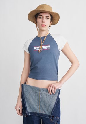 Noor inimene, kes kannab beežist mütsi ja sinist lühikest t-särki, hoides avatud siniste teksapükste vöökohta ühevärvilise tausta ees.