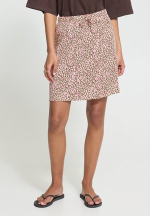 Femme portant une jupe beige et rose à imprimé léopard avec un cordon, un haut marron, des tongs noires, debout sur un fond uni.