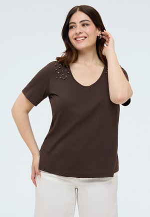 Mujer sonriendo, lleva una camiseta de manga corta de color marrón oscuro con detalles de tachuelas en los hombros y pantalones de color claro, de pie frente a un fondo liso.