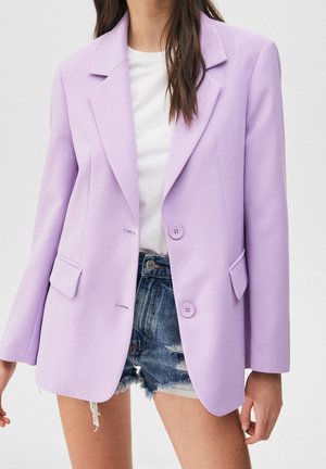 Femme portant un blazer violet clair sur un t-shirt blanc et un short en jean déchiré, debout devant un fond uni.
