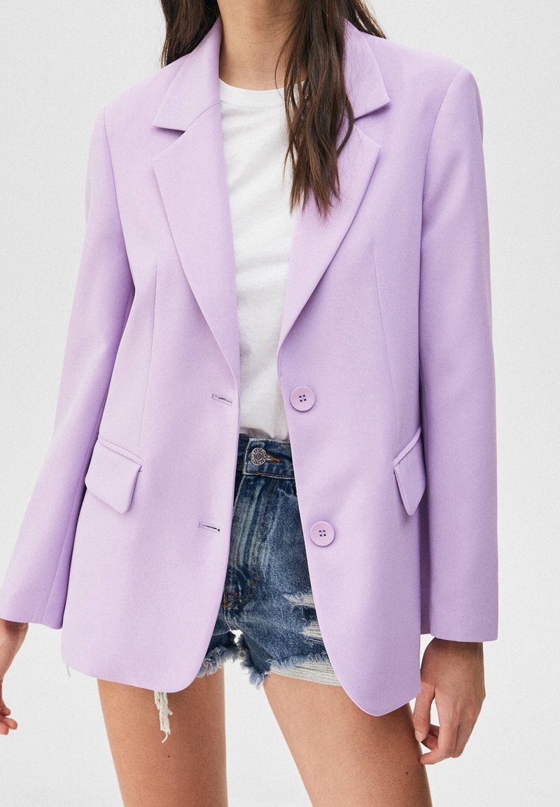 Femme portant un blazer violet clair sur un t-shirt blanc et un short en jean déchiré, debout devant un fond uni.