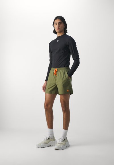Haut de sport à manches longues noir, shorts vert olive avec cordon orange, et baskets blanches, sur un fond clair uni.