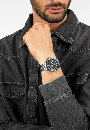Orologio in acciaio inox con quadrante nero, lunetta blu e bracciale a maglie argentate. Indossato al polso, abbinato a una camicia di jeans.