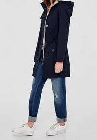 Navy waterproof parka with a hood, drawstring waist, button closures, and side pockets. Paired with rolled-cuff distressed jeans and white sneakers.