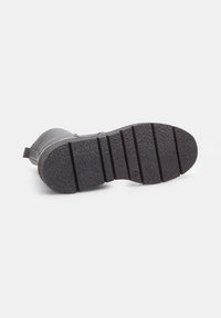 Suela de bota de goma gris con una superficie texturizada, que presenta surcos profundos para un mejor agarre y un leve tacón. Lengüeta trasera reforzada para facilitar el calzado.