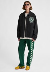 Svart knappad jacka med grön broderad logotyp, tillsammans med gröna träningsbyxor med vita chevronsjok och gula sneakers.