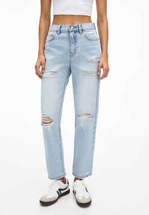 Person trägt hellblaue zerrissene Jeans, weißes Crop-Top und weiße Sneaker mit schwarzen und beigefarbenen Details, steht vor weißem Hintergrund.