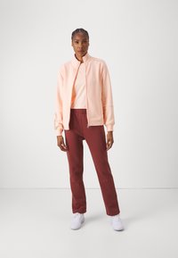 Sudadera de cierre zip en rosa claro, con puños acanalados, combinada con pantalones de chándal color burdeos, con cintura elástica, y zapatillas blancas. Diseño minimalista, ajuste casual.