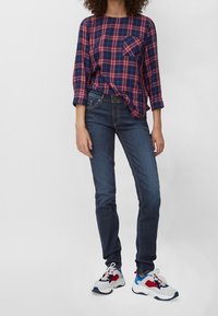Camisa de cuadros rojo y marino con un bolsillo, combinada con jeans oscuros ajustados de color azul y zapatillas blancas con detalles en rojo y azul.