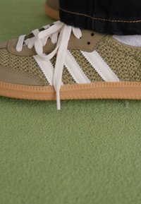 Sapatilha verde e bege com parte superior de tecido trançado, riscas brancas e sola de borracha natural. Apresenta atacadores redondos e um design texturizado na biqueira.