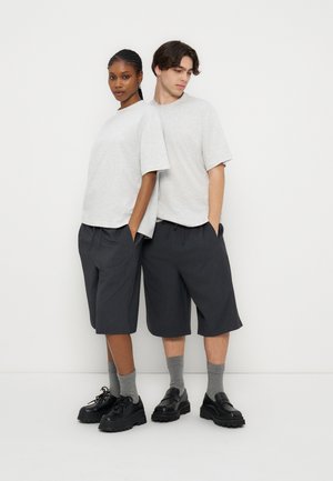 Dwóch modeli stoi tyłem do siebie, nosząc oversize jasnoszare t-shirty, czarne szorty do kolan, szare skarpety i czarne masywne buty.