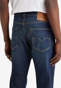 Jeans de mezclilla azul oscuro con dos bolsillos traseros, costuras amarillas contrastantes y un parche de cuero marrón en la cintura que muestra detalles de la marca.