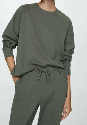 Personne portant un sweat-shirt ample vert olive à manches longues et un pantalon à cordon assorti, la main dans la poche, sur fond blanc.