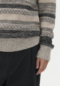 Beige och svart randig stickad tröja med ärm över svarta byxor, som visar en avslappnad hand vilande vid sidan mot en vit bakgrund.