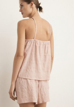 Mujer vistiendo una blusa sin mangas suelta de color rosa pálido con tirantes finos y pantalones cortos a juego, vista desde atrás sobre un fondo liso.