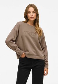 Brun sweatshirt med ordet "SANNING" broderat på framsidan, med en avslappnad passform och ribbade ärmar; kombinerad med svarta byxor.
