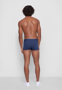 Marineblå boxershorts med myk elastisk linning, laget av et glatt stoff, sett fra baksiden på en modell som har på seg hvite sokker.
