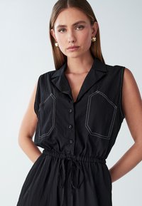 Mlada ženska z ravnimi lasmi nosi brezrokavno črno obleko s gumbi, belimi šivi na žepih in vrvico v pasu, pozira ob enobarvni ozadju.