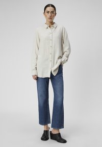 Chemise beige clair à boutons avec un tissu texturé, associée à un jean large bleu foncé. Le mannequin porte des mules noires à talons.