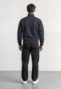 Man standing with back facing camera wearing a black jacket, black pants, and white sneakers on a gray floor against a white wall.