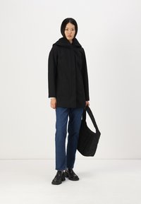 Veste à capuche noire au design épuré, avec deux poches avant et une texture lisse. Associée à un jean bleu et un sac noir.