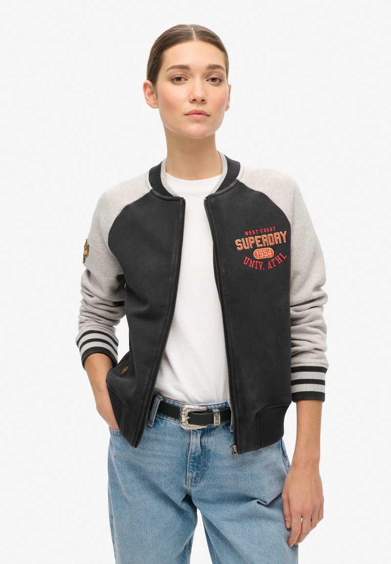 Veste bomber noire et grise avec fermeture éclair, poignets côtelés et logo rouge brodé. Présente une texture lisse et un design athlétique.