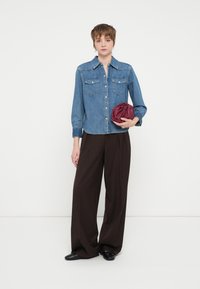 Jasnoniebieska koszula dżinsowa z guzikiem z przodu, kieszeniami na piersi i długimi rękawami, w zestawieniu z szerokimi brązowymi spodniami oraz zgniecioną burgundową torbą.