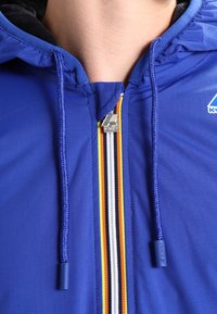 Blaue Kapuzenjacke mit Reißverschluss und gestreiftem Muster in Gelb, Weiß, Orange und Schwarz. Silberne Beschläge mit Logodetail.