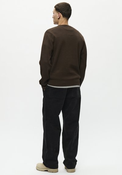 Suéter marrón con textura y cuello redondo, combinado con pantalones de pana negros. Modelo con calzado claro posando frente a un fondo liso.