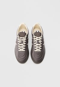 Ζευγάρι σκούρων καφέ αθλητικών παπουτσιών με μπεζ κορδόνια και ραφές, ετικέτες με την επωνυμία στη γλώσσα της γλώσσας και λαστιχένιες σόλες σε λευκό φόντο.