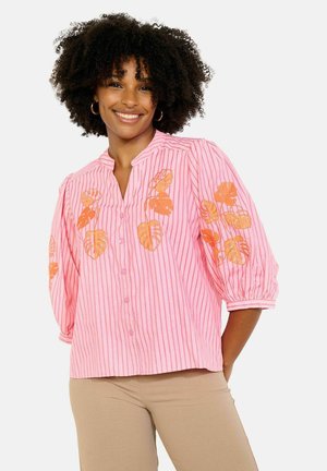 Uśmiechnięta kobieta o kręconych włosach, ubrana w różową bluzkę w paski z pomarańczową haftowaną aplikacją liści oraz beżowe spodnie, stoi z rękami za plecami.