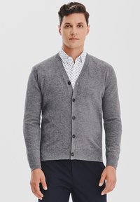 Mężczyzna w szarej kardiganie na guziki, noszący biał�ą koszulę w wzory i ciemne spodnie, stojący na tle jasnym.