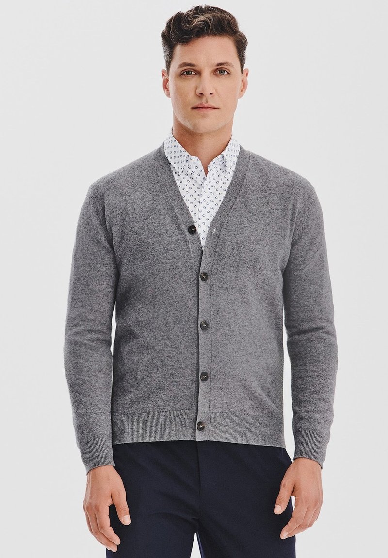 Mężczyzna w szarej kardiganie na guziki, noszący białą koszulę w wzory i ciemne spodnie, stojący na tle jasnym.